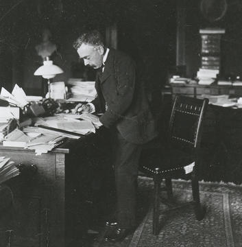 Black-and-white photograph of William Gilson Farlow standing at a desk covered with papers and books, writing with a pen, circa 1900