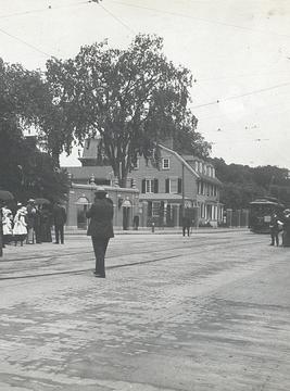 Black-and-white photograph of Harvard Square in 1903, showing people walking on a dirt road with trees and wooden buildings in the background