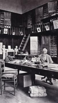 Black-and-white photograph of Sereno Watson seated at a large desk surrounded by books, papers, and specimens in the Gray Herbarium at Harvard, dated 1880