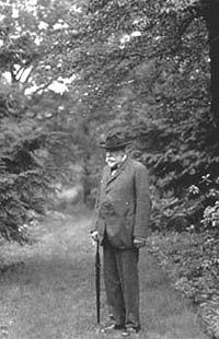 Black-and-white photograph of Charles Sprague Sargent standing on a tree-lined path at the Arnold Arboretum, holding a walking stick and wearing a dark suit and hat
