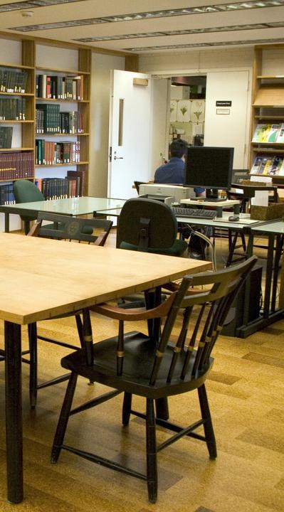 Interior view of the library reading room with bookshelves, tables, chairs, desktop computers, and a person seated at a computer near the back wall