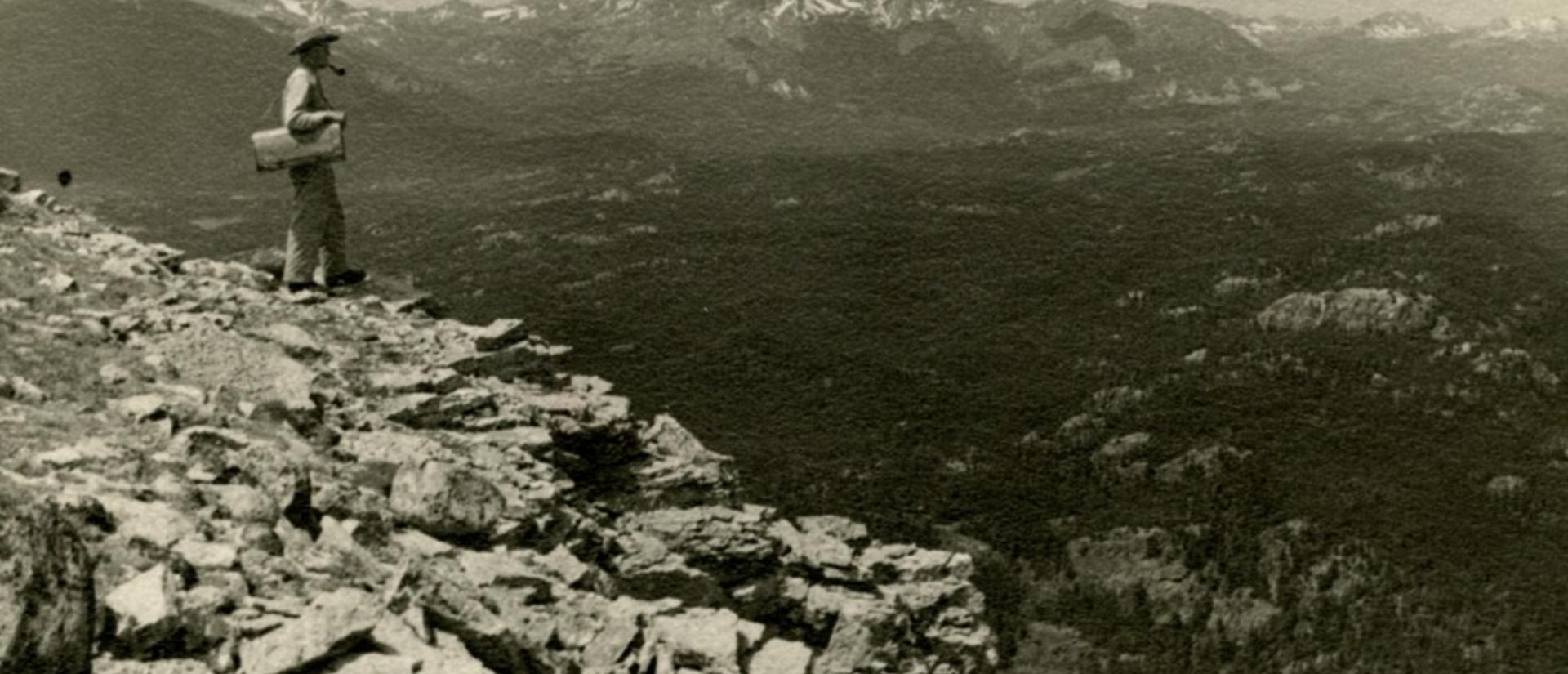 Black-and-white photograph of Reed Rollins standing on a rocky slope overlooking a mountainous landscape with forested valleys in Wyoming, 1951