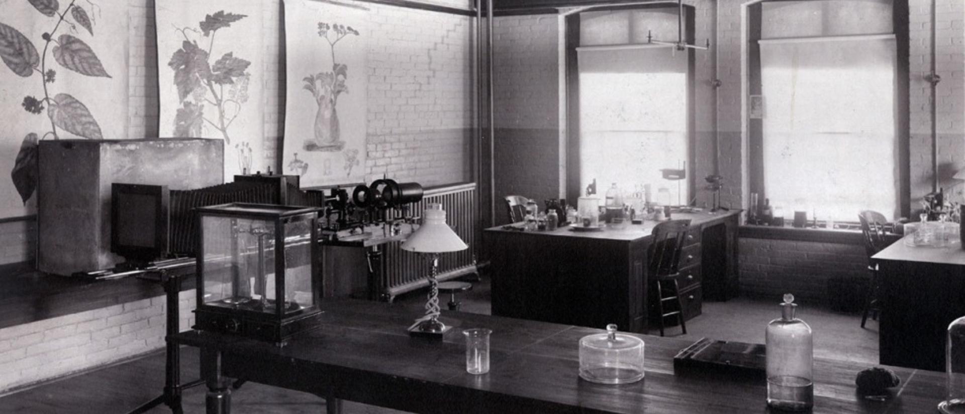 Black-and-white photograph of a botany classroom with tables, glass containers, botanical posters on the walls, and large windows in the University Museum building, circa 1900